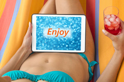 Midsection of young woman wearing bikini using digital tablet while resting on towel outdoors