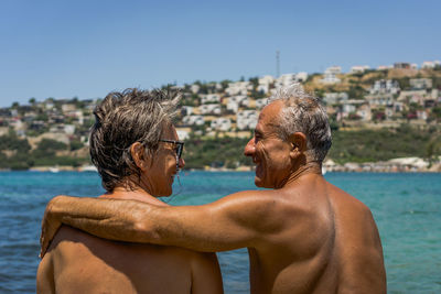 Rear view of shirtless couple standing at beach against clear sky