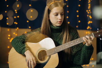 Beautiful teenage girl in christmas antlers and sweater plays guitar