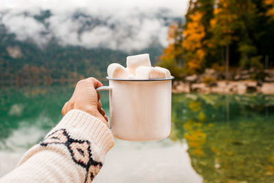 Picnic in bavarian mountains, germany. hand with mug of cacao and marshmallow on the lake background