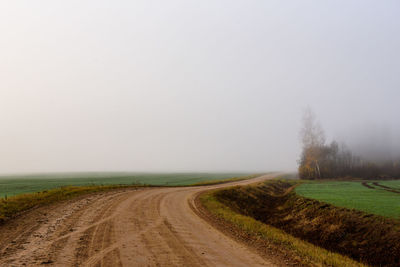 Dirt road amidst field against sky during foggy weather