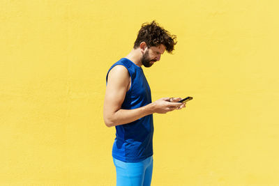 Side view of young man using mobile phone against yellow wall