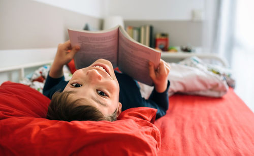 Portrait of smiling boy with book lying on bed at home