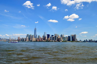 One world trade center amidst buildings in front of river against sky on sunny day