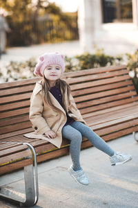 Stylish kid girl 3-4 year old wearing trendy autumn coat and knitted hat sittin on bench in park