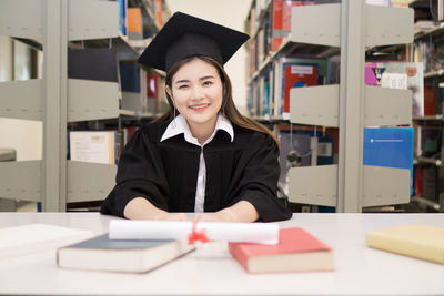 Smiling young woman in graduation gown sitting at table in library