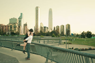 Side view of boyfriend carrying girlfriend by railing against cityscape