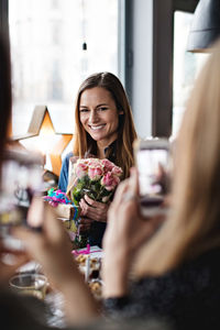 Female friends photographing smiling woman with bouquet at dining table in restaurant