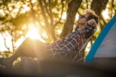 Side view of woman sitting on chair against trees during sunset