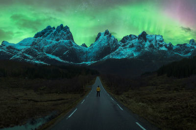 Back view of anonymous traveler in yellow jacket standing on asphalt road and admiring spectacular view of huge rocky mountains with snowy peaks under sky with polar lights in lofoten person