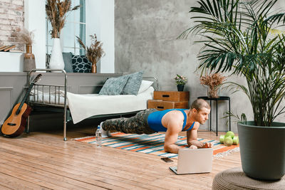 Cheerful sportsman with black hair makes plank and watching online workout from laptop in bedroom