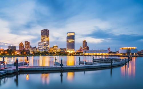 Illuminated buildings by river against sky at dusk