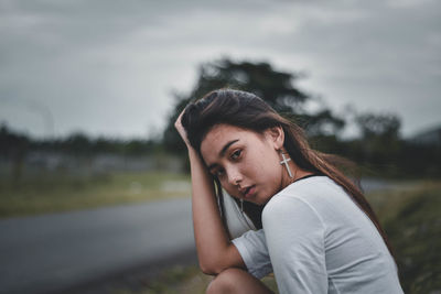 Portrait of young woman with hand in hair sitting against sky