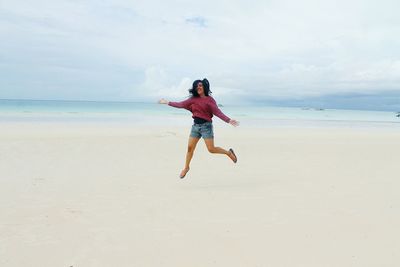 Full length of young woman jumping at beach against sky