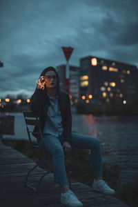 Portrait of young woman sitting on chair at riverbank in city during dusk