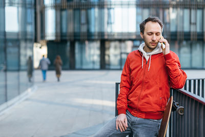 Close up portrait of bearded trendy man in red jacket and jeans. man is talking on the mobile phone.
