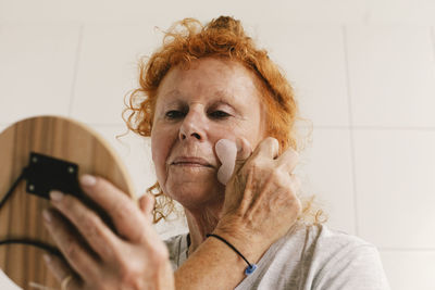 Senior woman looking in mirror and massaging face against wall