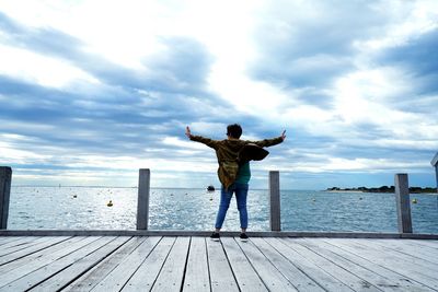 Rear view of carefree man with arms outstretched standing on pier by sea against cloudy sky