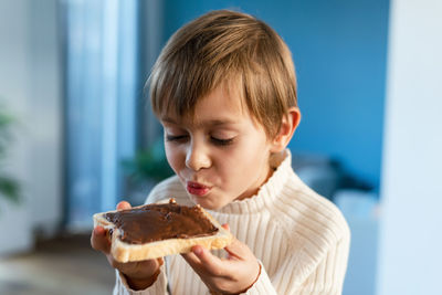A boy bites into a sandwich with hazelnuts and cocoa, sitting on a table in the living room.