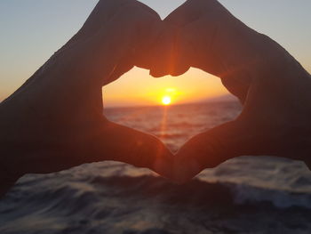 Cropped hands making heart shape against sea during sunset