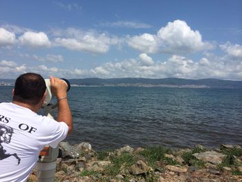 Rear view of man looking through coin-operated binoculars at beach against cloudy sky