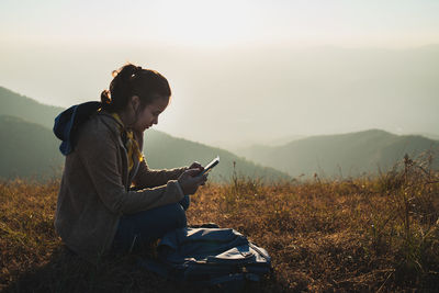 Side view of asian female traveler sitting on the top of the hill and using a mobile phone