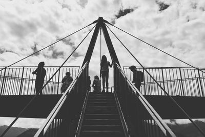 Low angle view of people on footbridge against cloudy sky