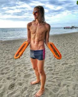 Full length of shirtless man holding papaya at beach against sky