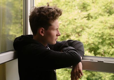 Side view of thoughtful young man looking through window at home