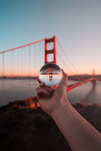 Person hand holding bridge against sky during sunset