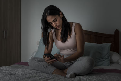 Young woman using phone while sitting on bed at home