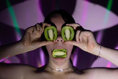 Close-up of young woman covering face with kiwi fruit against wall