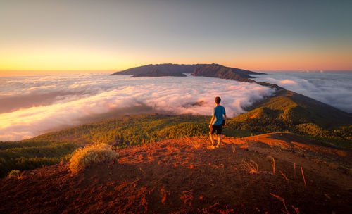 Back view traveler standing on hill while admiring spectacular scenery on clouds over mountainous terrain