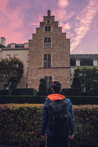 Rear view of man with backpack looking at building against dramatic sky