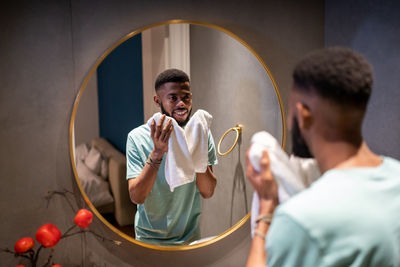 Pleased african american man looks in mirror holds bath towel smiling enjoy good morning before work