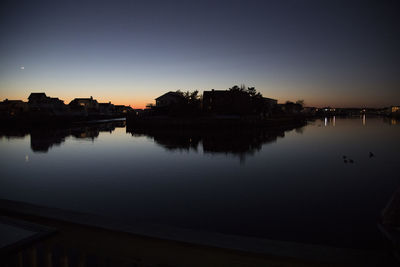 Silhouette buildings by lake against sky during sunset in city