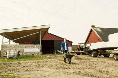 Man pushing wheelbarrow on dirt road by tractor against barn at farm