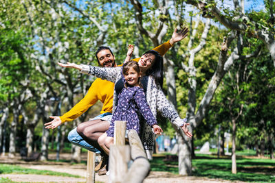 Portrait of smiling parents and daughter with arms outstretched sitting on railing against trees in park
