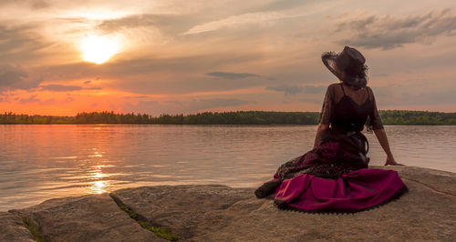 Woman sitting by lake against sky during sunset