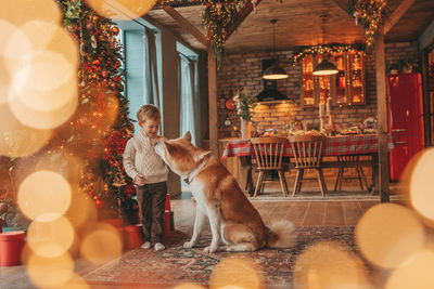 Candid authentic happy little boy in knitted beige sweater hugs dog with bow tie at home on xmas