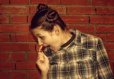 Young woman holding lollipop while standing against brick wall