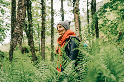 Young active woman hiker in a jacket and a hat with a backpack in a thicket of ferns in the forest