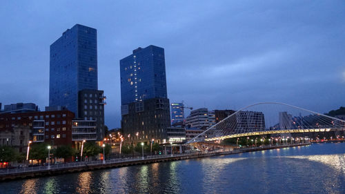 Illuminated buildings by river against sky in city at dusk