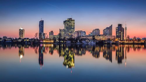 Illuminated cityscape by river against clear sky during sunset