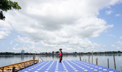 Woman standing on pier by lake against cloudy sky