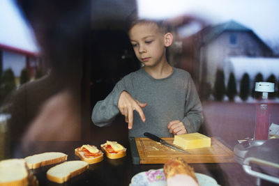Boy preparing sandwich at table seen through glass window