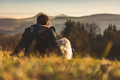 Rear view of man with dog sitting on field against sky