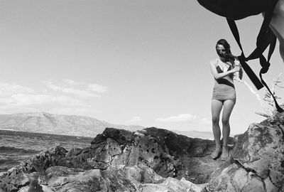 Fill length of young woman standing on rock at beach against sky