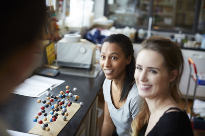 Smiling young female students by molecular structure looking at mature teacher in chemistry classroom