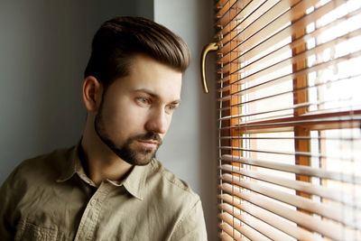 Close-up of thoughtful young man looking through window blinds